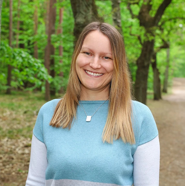 Melli Seedorf, sehr freundlich lächelnde Glückstrainerin, steht auf einem grünen Waldweg